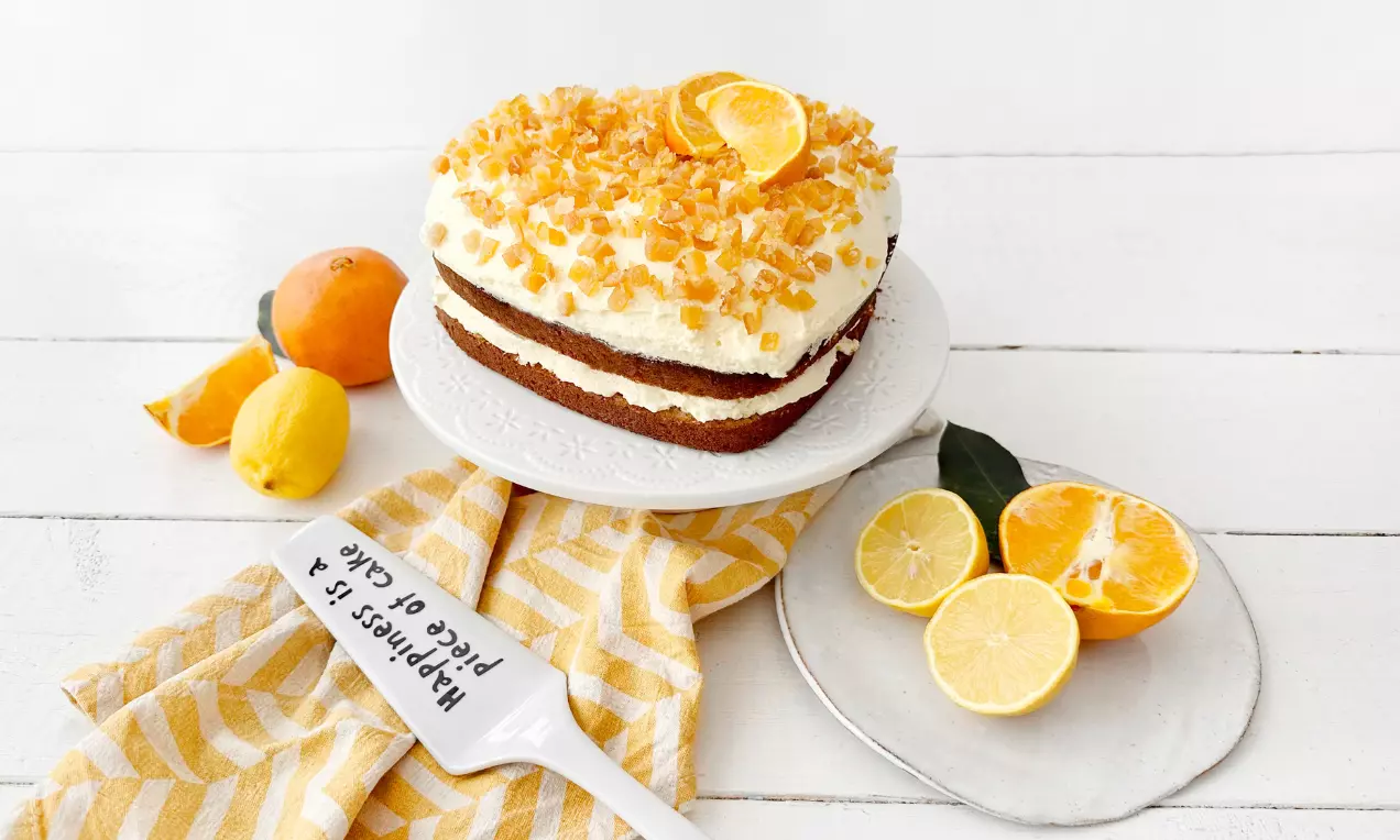 Ambrosiatårta med två gyllenbruna bottnar, vit glasyr, apelsinskal och kanderade apelsinbitar på vit tallrik med citrusfrukter och randig duk