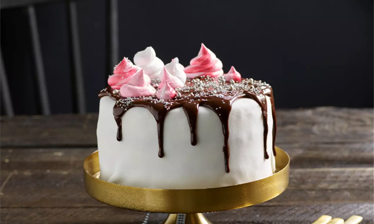 Vit täckt tårta med blank chokladglasyr som droppar ner på sidorna, dekorerad med rosa och vita maränger på ett guldigt tårtfat