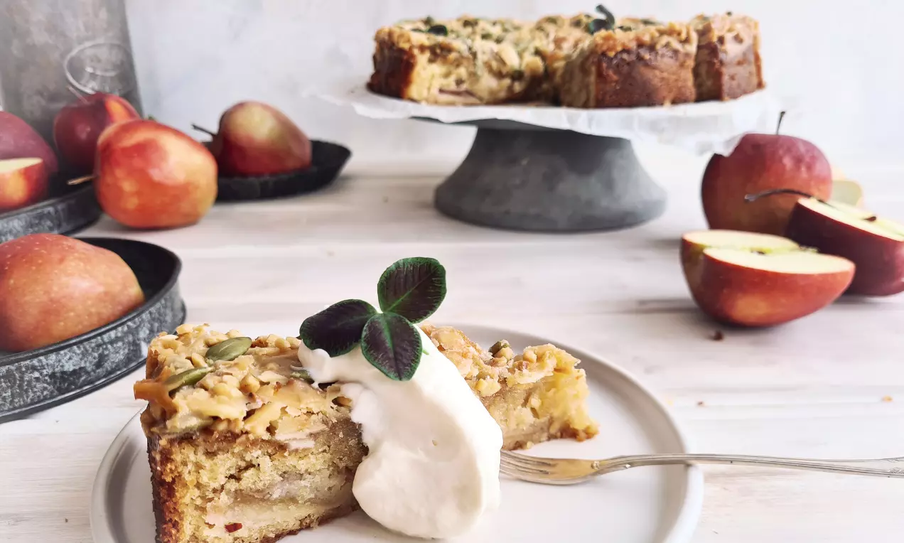 Äppelkaka med gyllenbrunt toscatäcke serverad med grädde på en vit tallrik, dekorerad med gröna blad och omgiven av röda äpplen