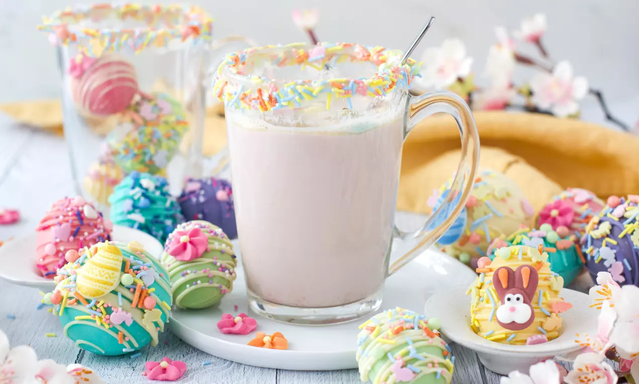 Färgglada chokladägg med dekorativa strössel och mönster på en vit tallrik bredvid glas med kant av strössel och ljus dryck