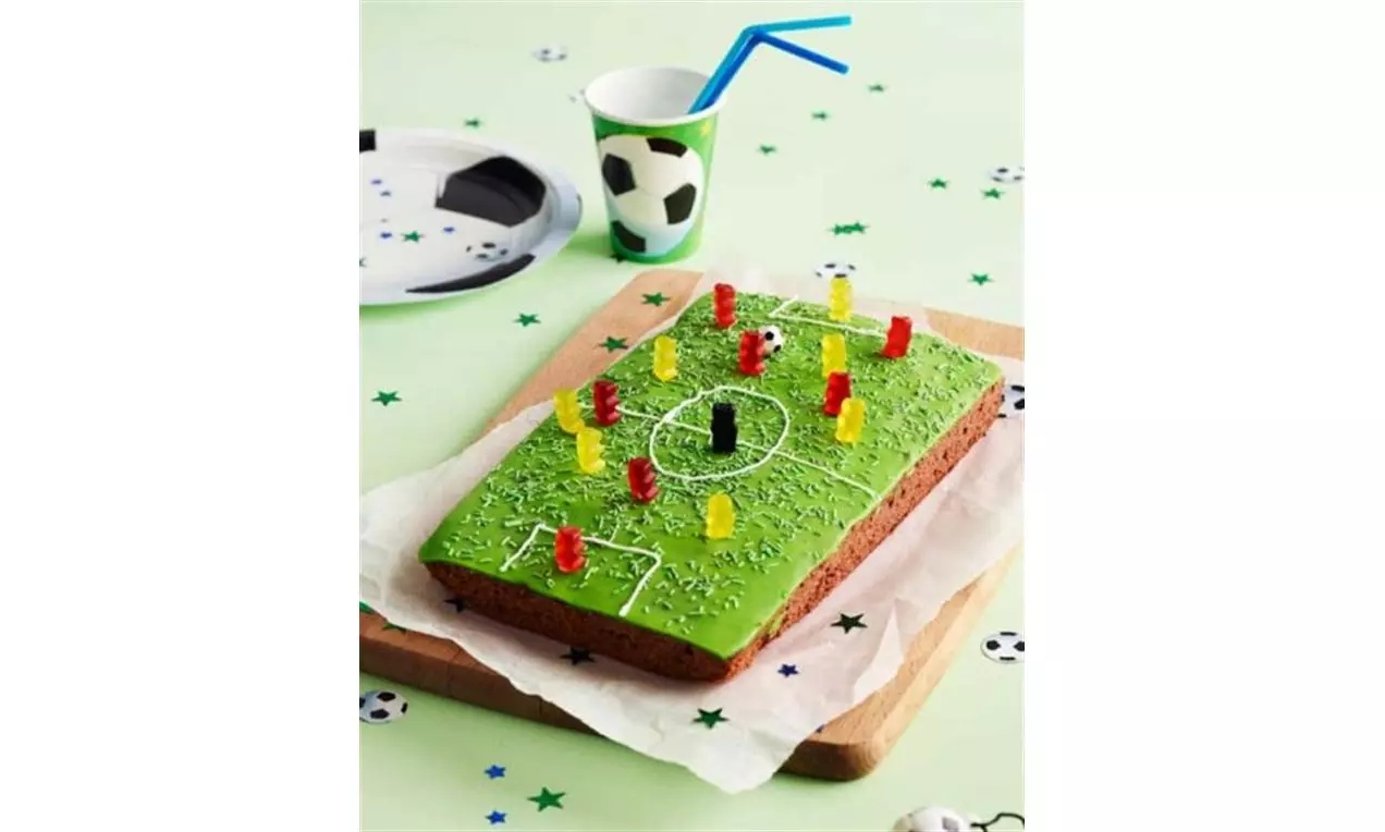 Rektangulär fotbollstårta med grön glasyr, vita linjer som fotbollsplan, dekorerad med stående gelégodisfigurer, på träbricka med papper