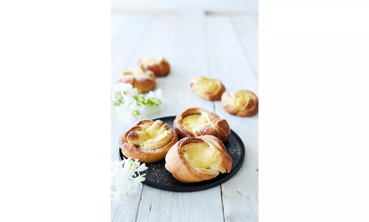 Vaniljkrämbullar med gyllene spiralformad deg och ljusgul kräm på svart tallrik, dekorerad med vita blommor mot ljust träbord
