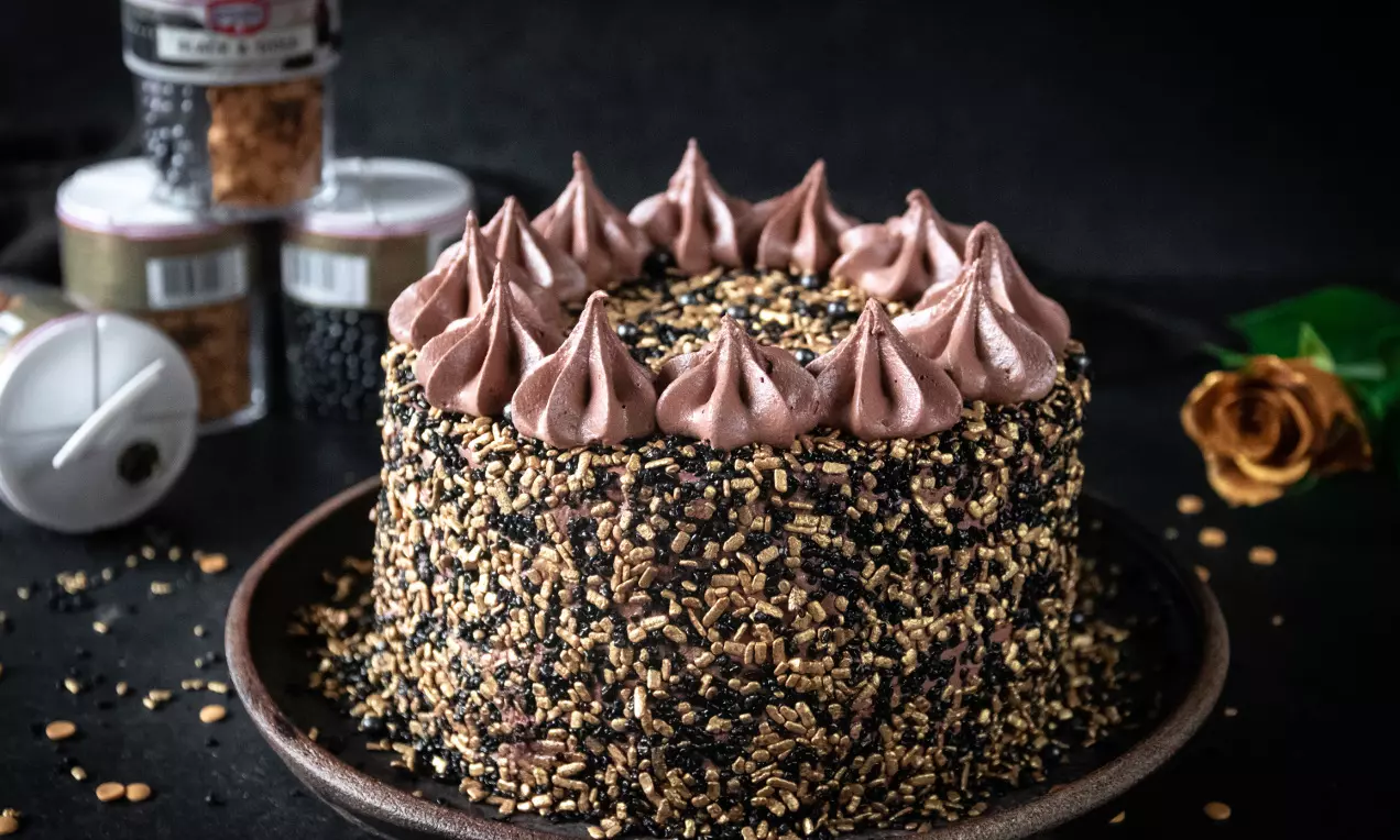 Nyårstårta med chokladmousse-toppar, täckt av svart och guldströssel på brun tallrik, omgiven av strösselburkar och en gyllene ros
