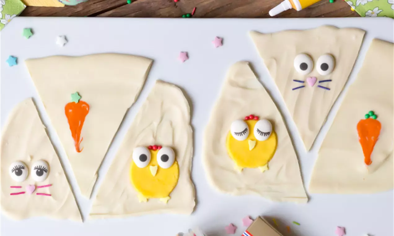 Vit choklad dekorerad som kycklingar och morötter med gula och orangea detaljer, placerad på en vit bricka omgiven av påskägg och dekorationer