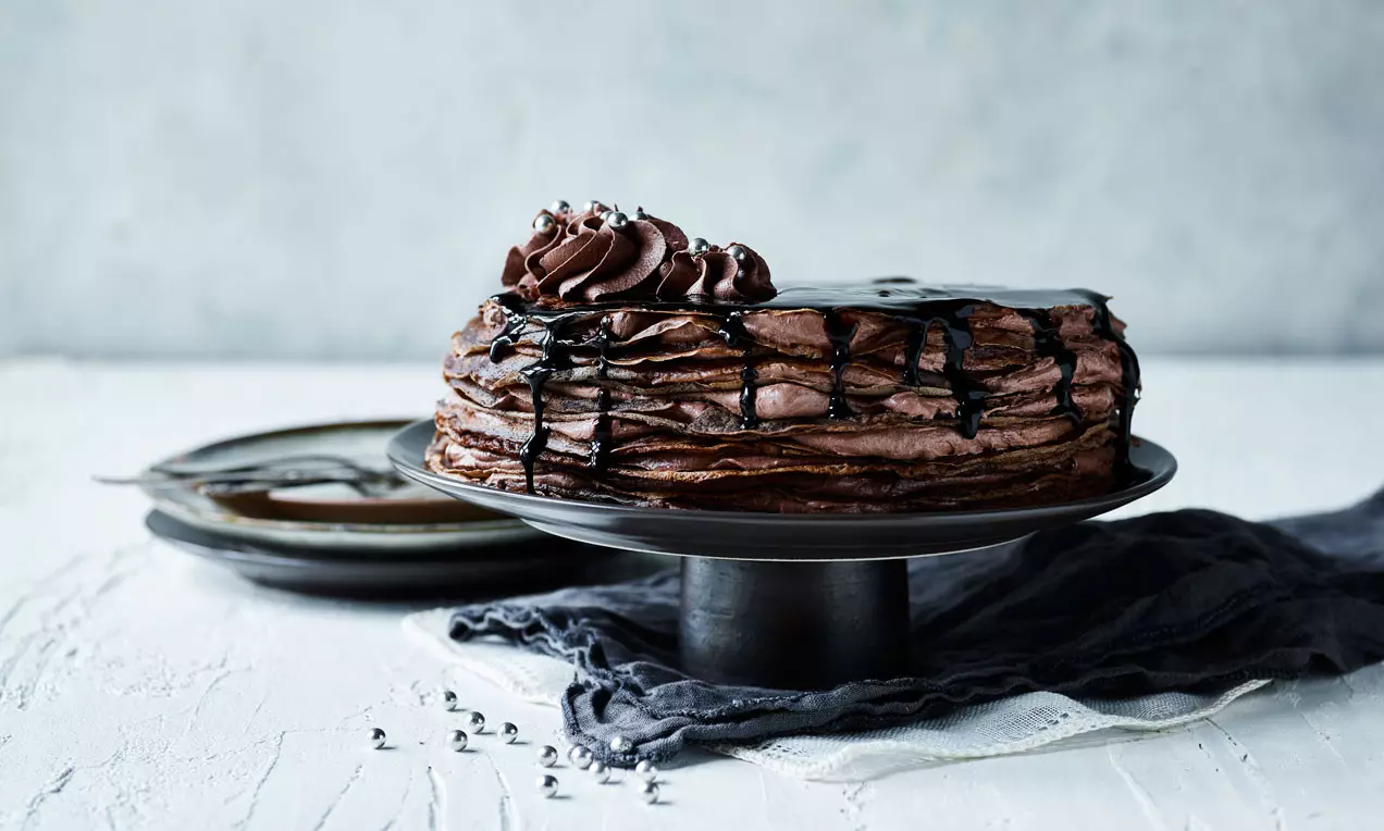 Pannkakstårta med lager av mörk chokladmousse, toppad med chokladsås och spritsade chokladrosetter på ett metallfat med mörk duk