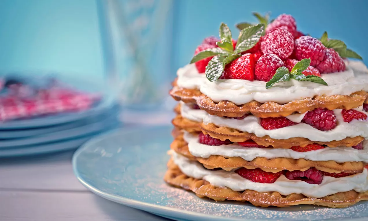 Naked cake med gyllene våfflor i lager, fylld med vit grädde och röda hallon, toppad med mynta och pudrad med florsocker på blå tallrik