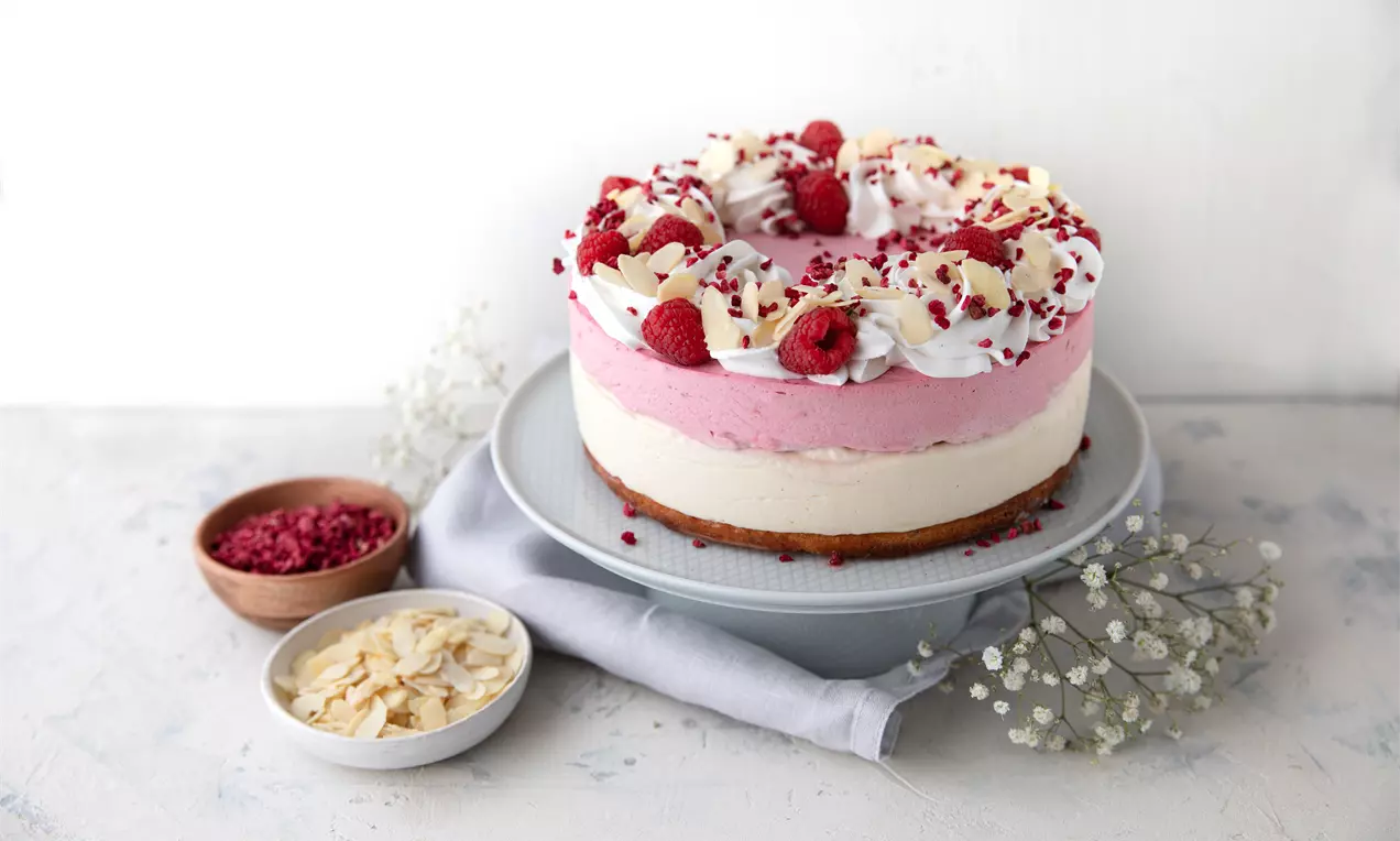 Semmeltårta med lager av vit och rosa fyllning, toppad med vispgrädde, mandelflarn, hallon och lingon på ett vitt tårtfat med dekorationer