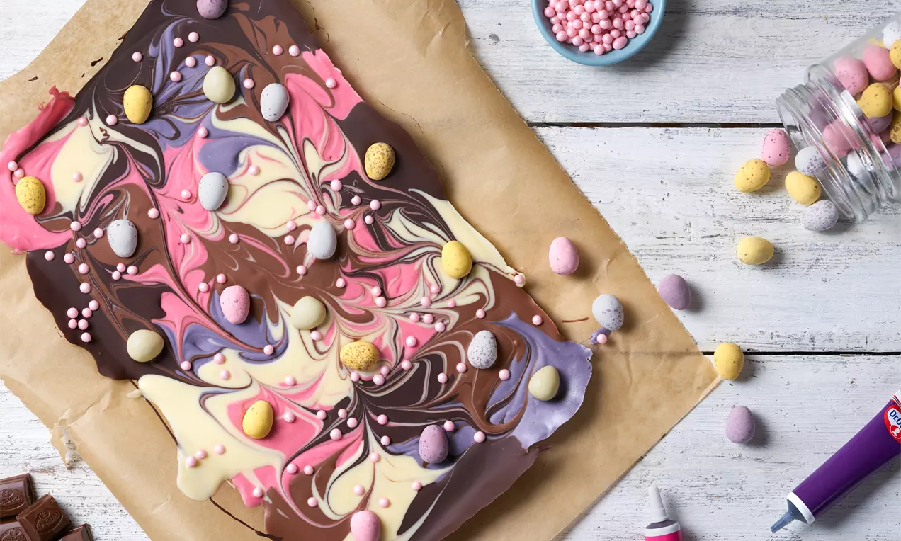 Chokladbräck med marmorering i brun, vit, rosa och lila, dekorerad med små ägg och pärlor på bakplåtspapper på ljus träyta