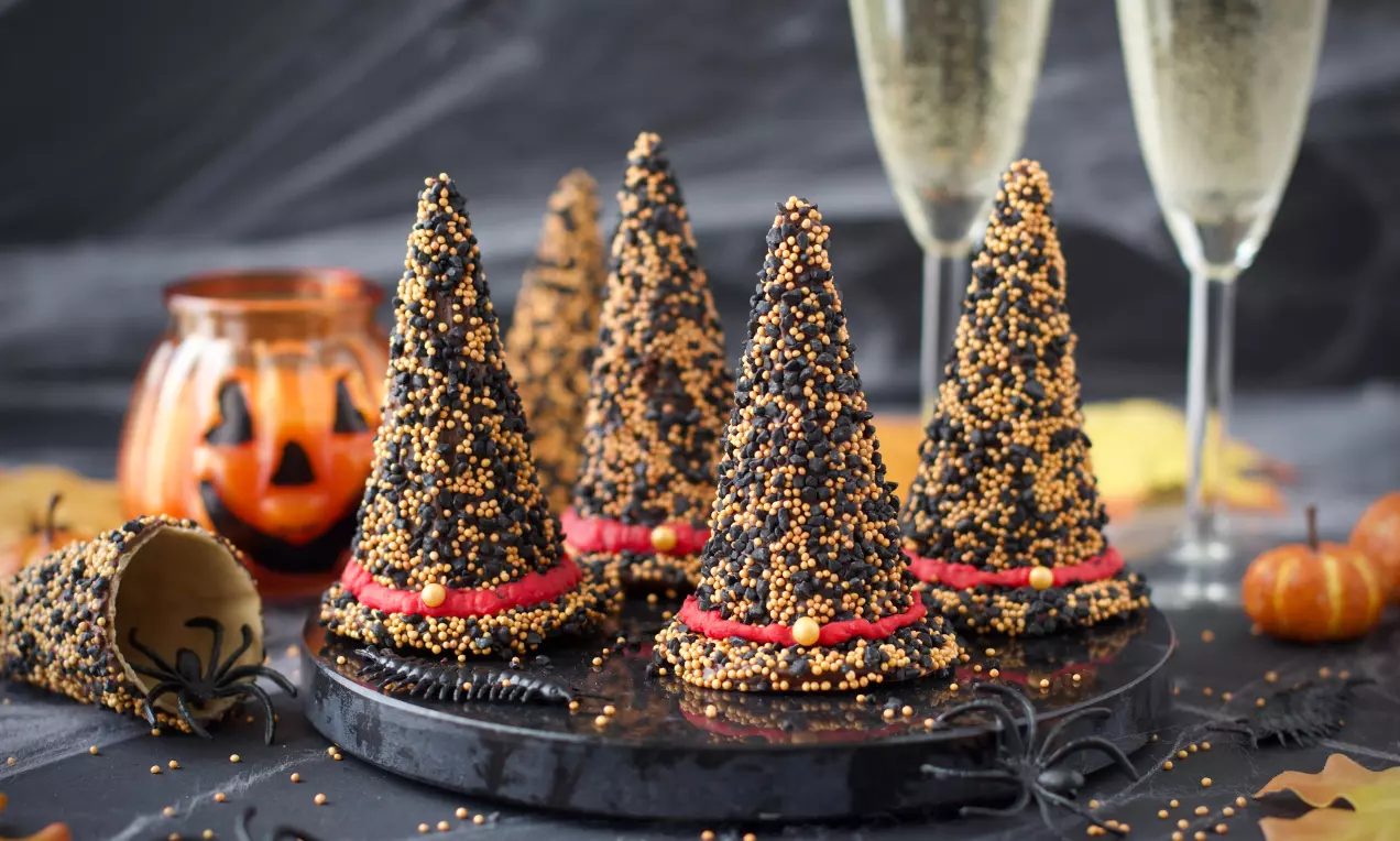 Höga svarta häxhattar täckta med gulddragéer och röda dekorband på svart bricka, omgivna av orange pumpor och glimmande champagneglas