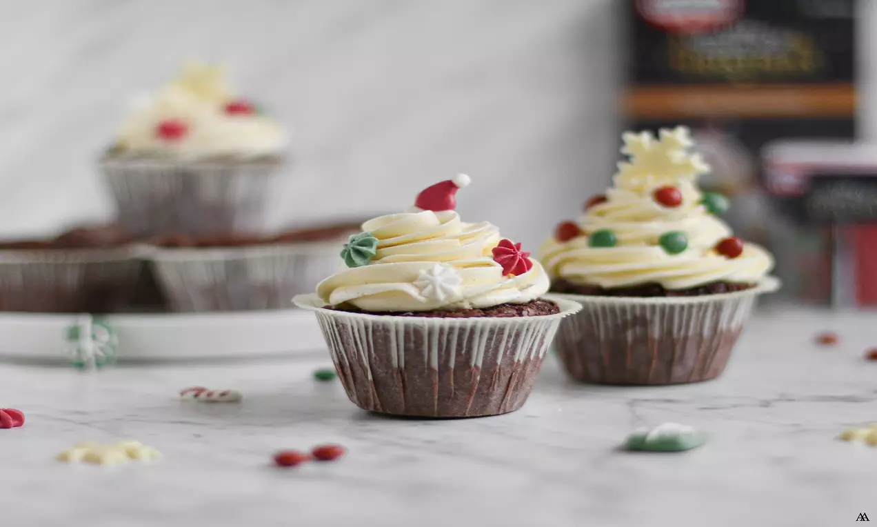 Chokladmuffins med krusig vit marängsmörkräm, dekorerade med färgglada julmotiv, placerade på marmorbord med strödda godisbitar