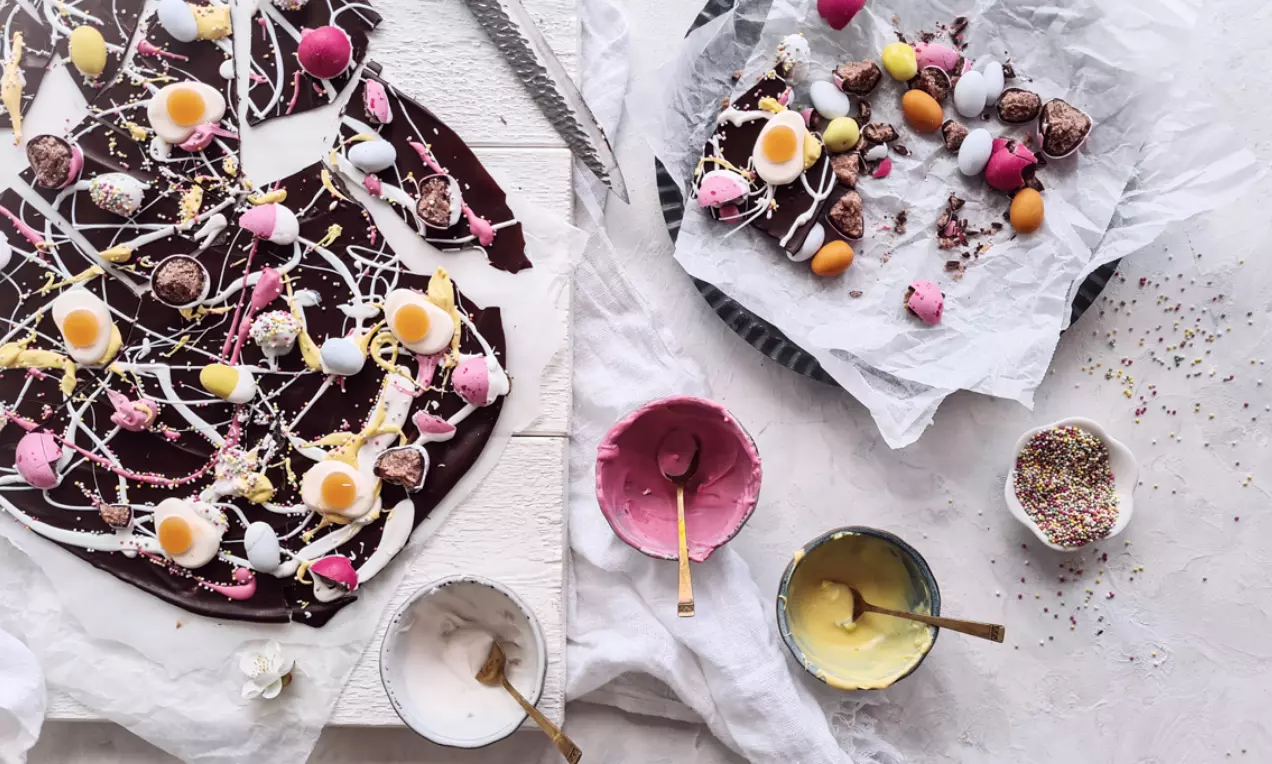 Mörk chokladbräck med vita och rosa chokladstreck, dekorerat med äggformade godisar och strössel, på bakplåtspapper med små skålar intill