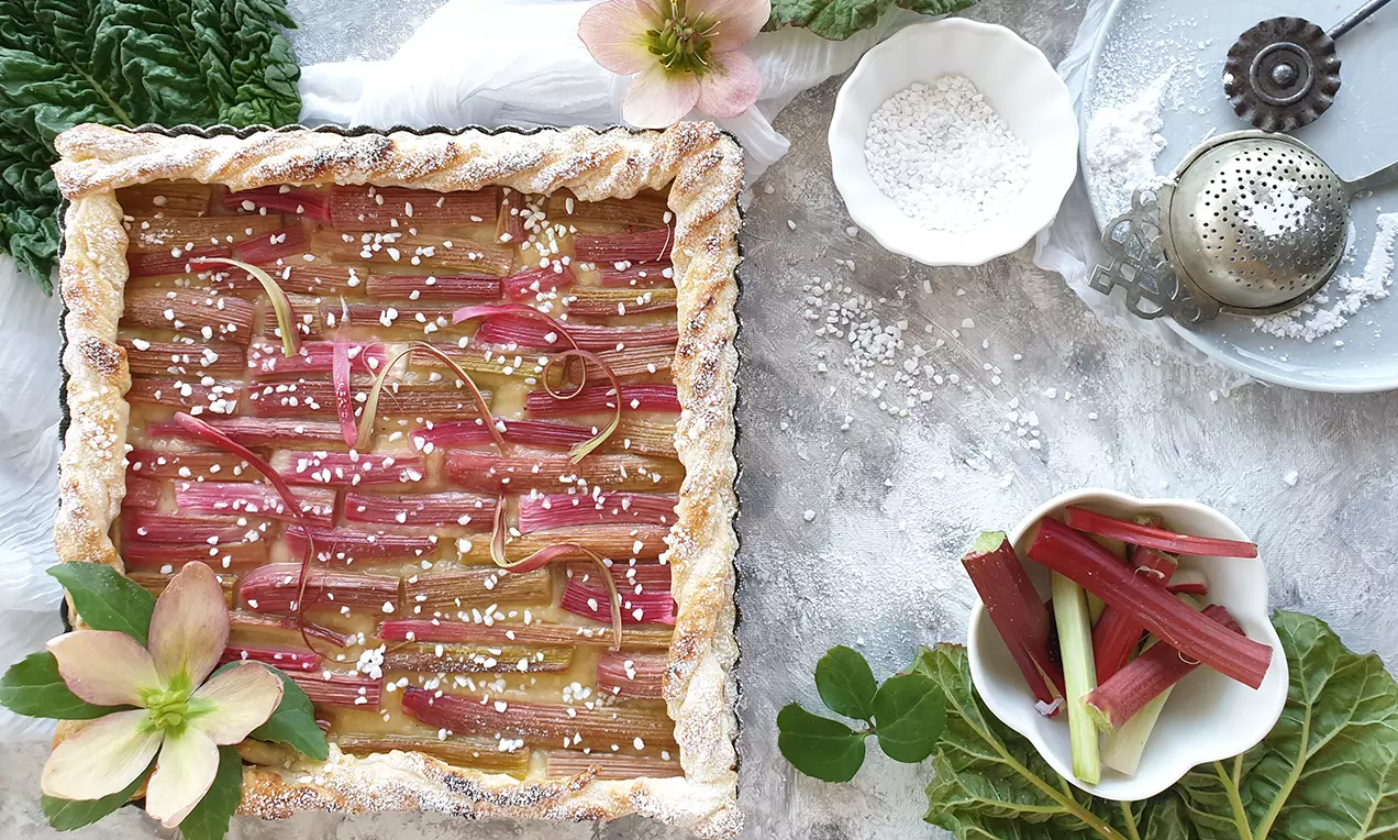 Rektangulär smördegspaj med gyllenbruna kanter, toppad med vaniljkräm och rabarber i röda och rosa strimlor, pudrad med vitt socker