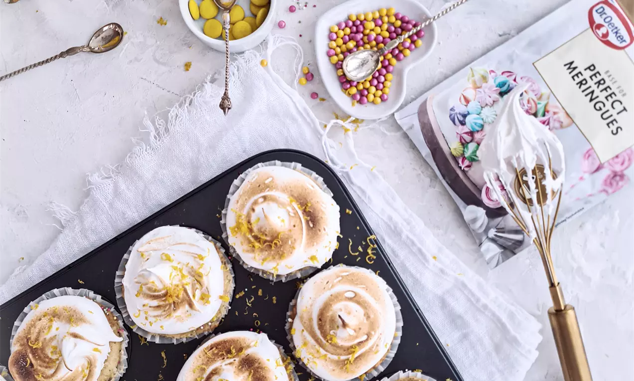 Citronmuffins med gyllene marängtoppar, rivet citronskal och färgglada strössel bredvid skål med citroner och dekorationer på ljus bakgrund