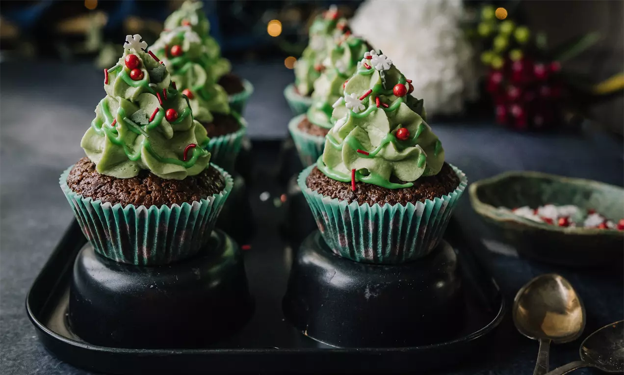 Chokladmuffins i blå-gröna formar toppade med grön grädde formad som granar med röda och vita dekorationer på mörk bricka