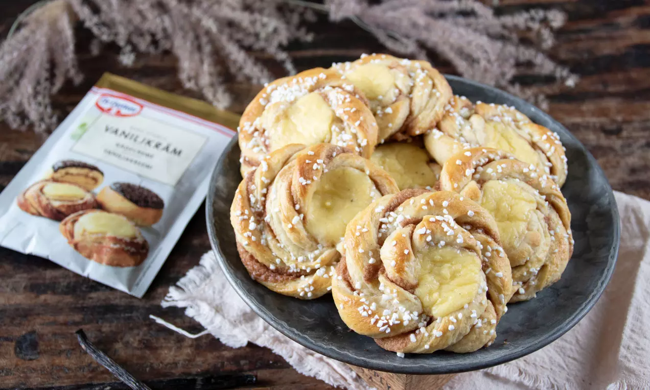 Kanelbullar gyllenbruna och flätade med en krämig ljusgul vaniljfyllning, strödda med vit pärlsocker på ett rustikt fat med beige duk