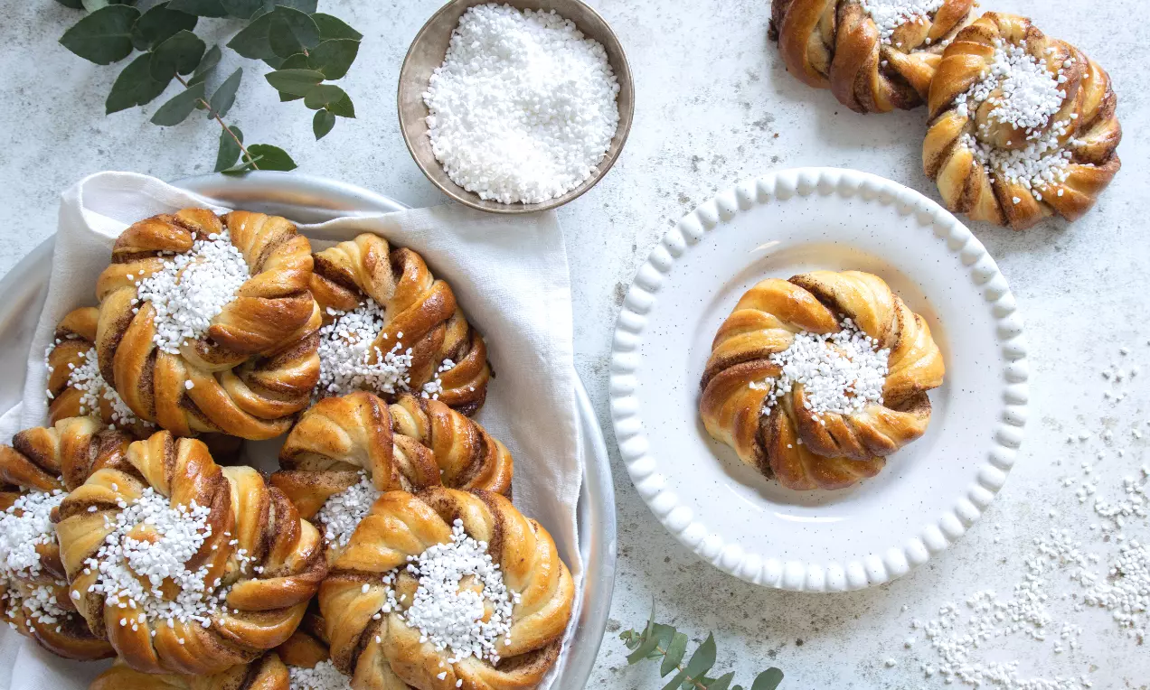 Gylenbruna kanelbullar med vriden form, täckta av pärlsocker, serverade på fat med enskild bulle upplagd på ett vitt tallriksunderlägg