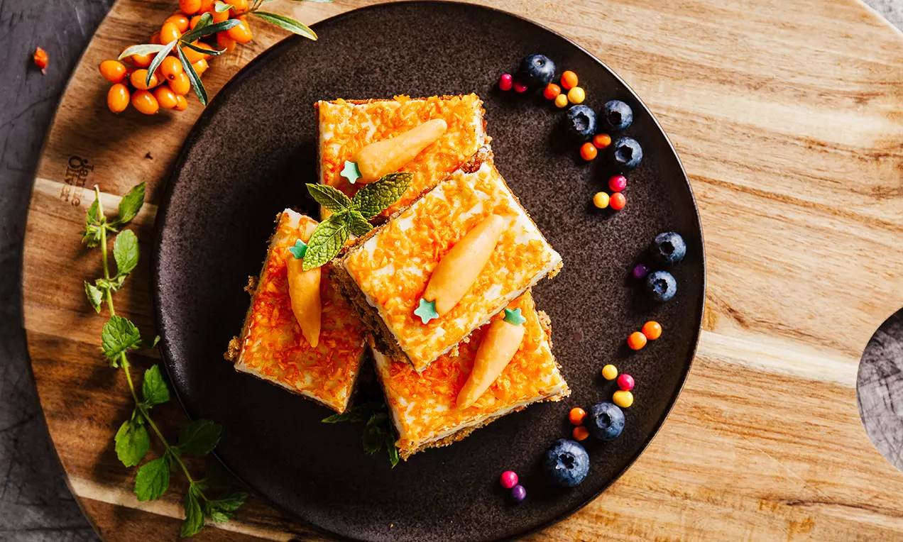 Fyrkantiga morotskakebitar med orange glasyr, pistagenötter och gröna myntablad, upplagda på mörk tallrik med bär och örtkvistar
