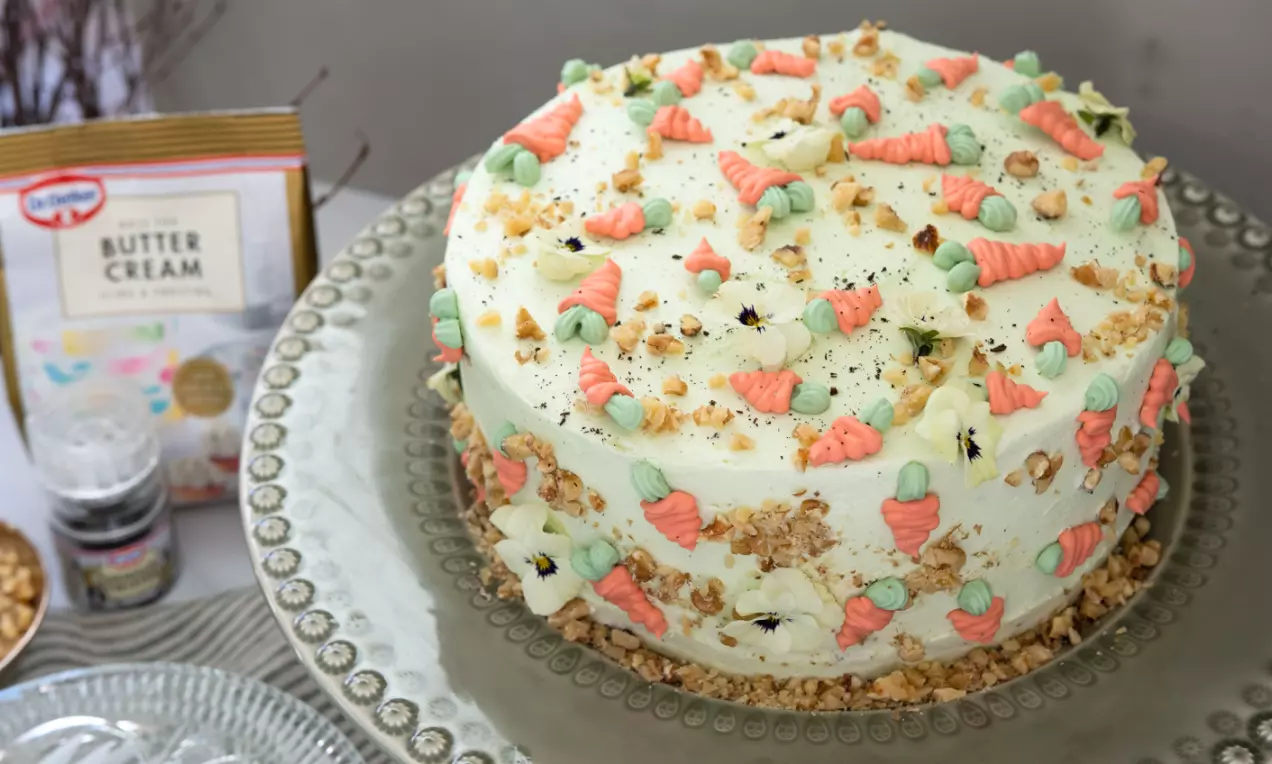Morotstårta med vit frosting, dekorerad med små kristyrmorötter i orange och grönt, hackade nötter och ett tunt smulskikt på fat
