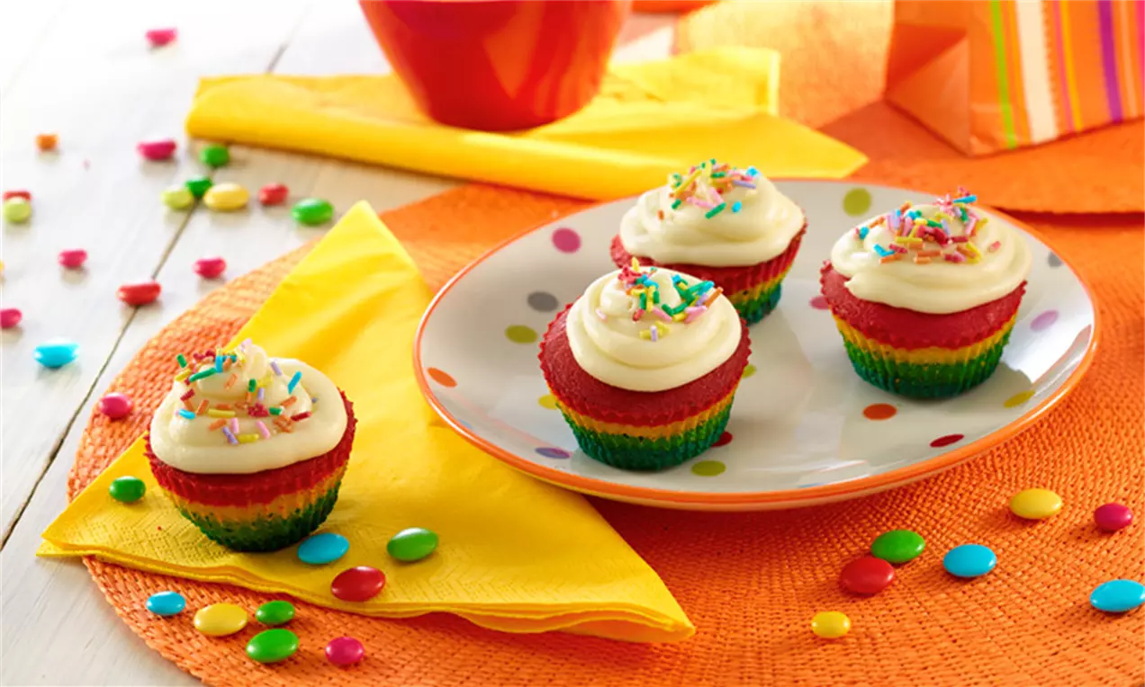 Fyra regnbågsfärgade cupcakes med vit frosting och strössel på en vit tallrik med prickar, omgivna av färgglada dekorationer