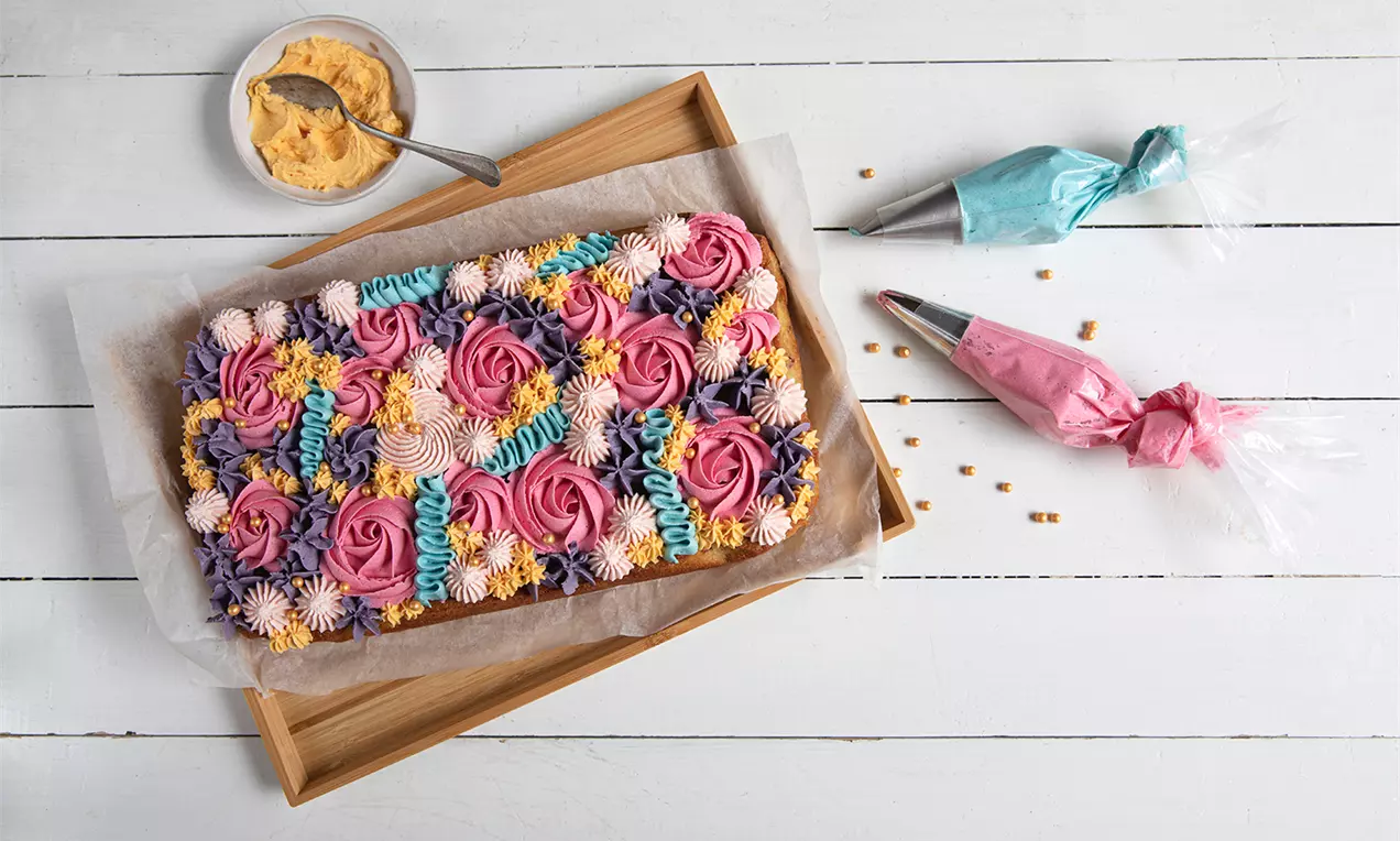 Långpannekaka dekorerad med färgglada spritsade rosor och blommor i rosa, lila, blått och gult på ett bakplåtspapper i en trälåda