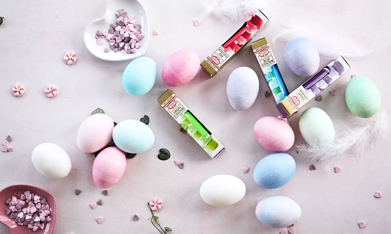 Pastellfärgade ägg i rosa, blått, grönt och lila på ljus yta, omgivna av gelfärgstuber, vita fjädrar och små dekorativa hjärtan