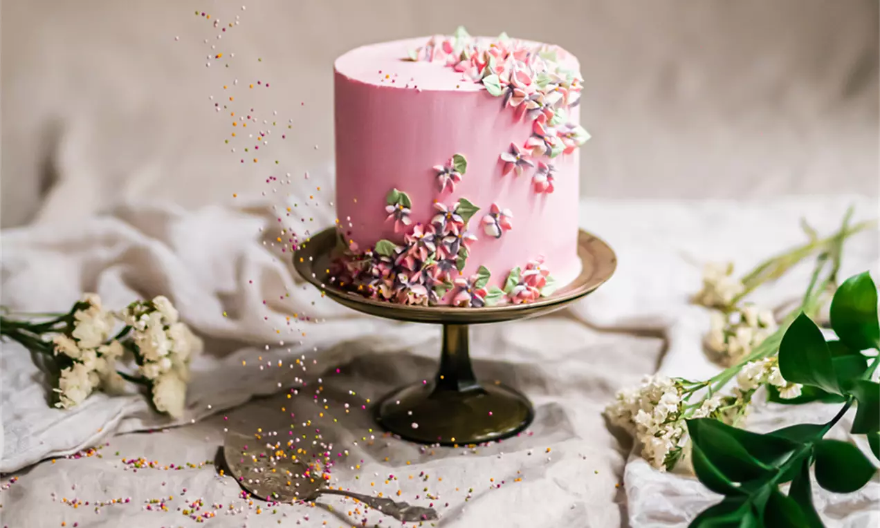 Hortensiatårta med rosa glasyr, dekorerad med små blommor i vitt och rosa, på en mörk tårtställning med gröna blad och vita blommor runtom