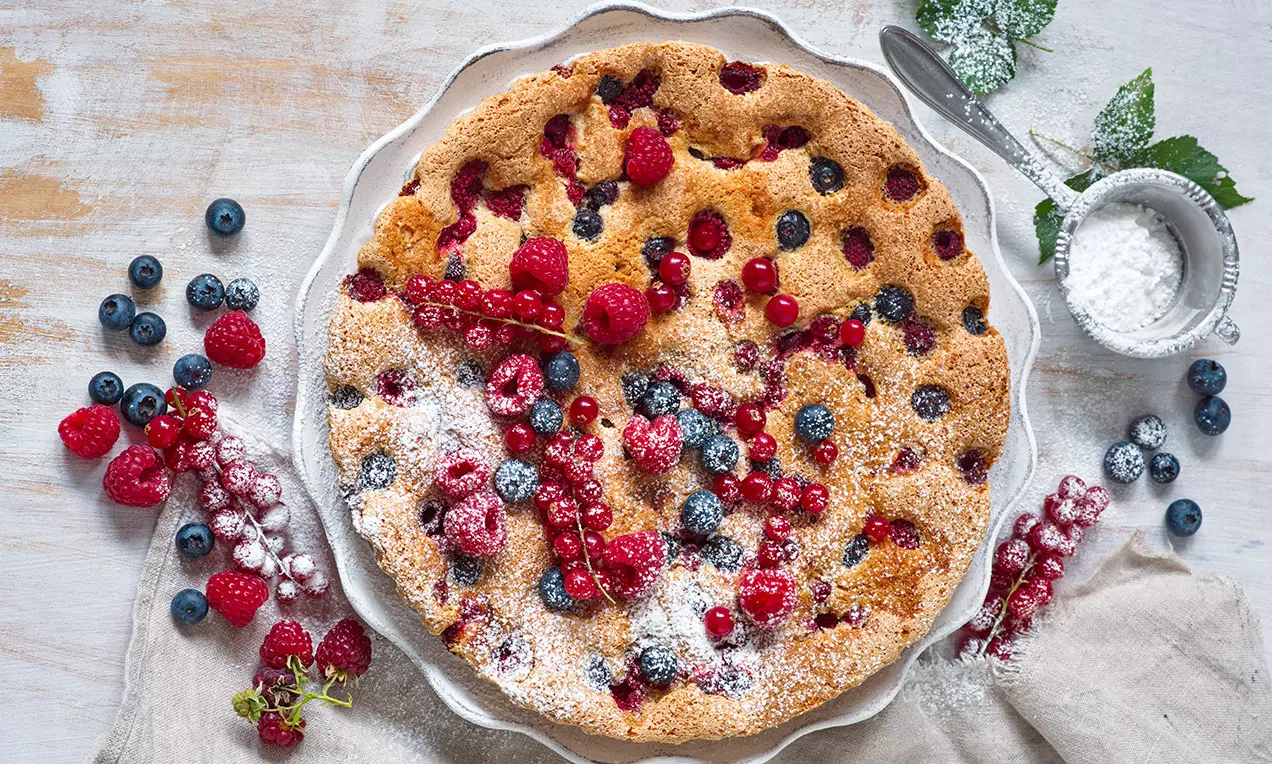 Somrig bärkaka med gyllenbrun, mjuk yta toppad med hallon, blåbär och röda vinbär, pudrad med florsocker och serverad på ett vitt fat