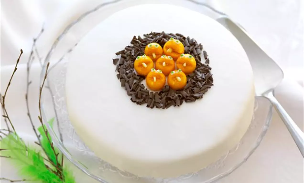 Moussetårta med vit slät yta, toppad med cirkel av chokladspån och orangefärgade bakverk, på glasfat med grönt fjädersmyckat ris.