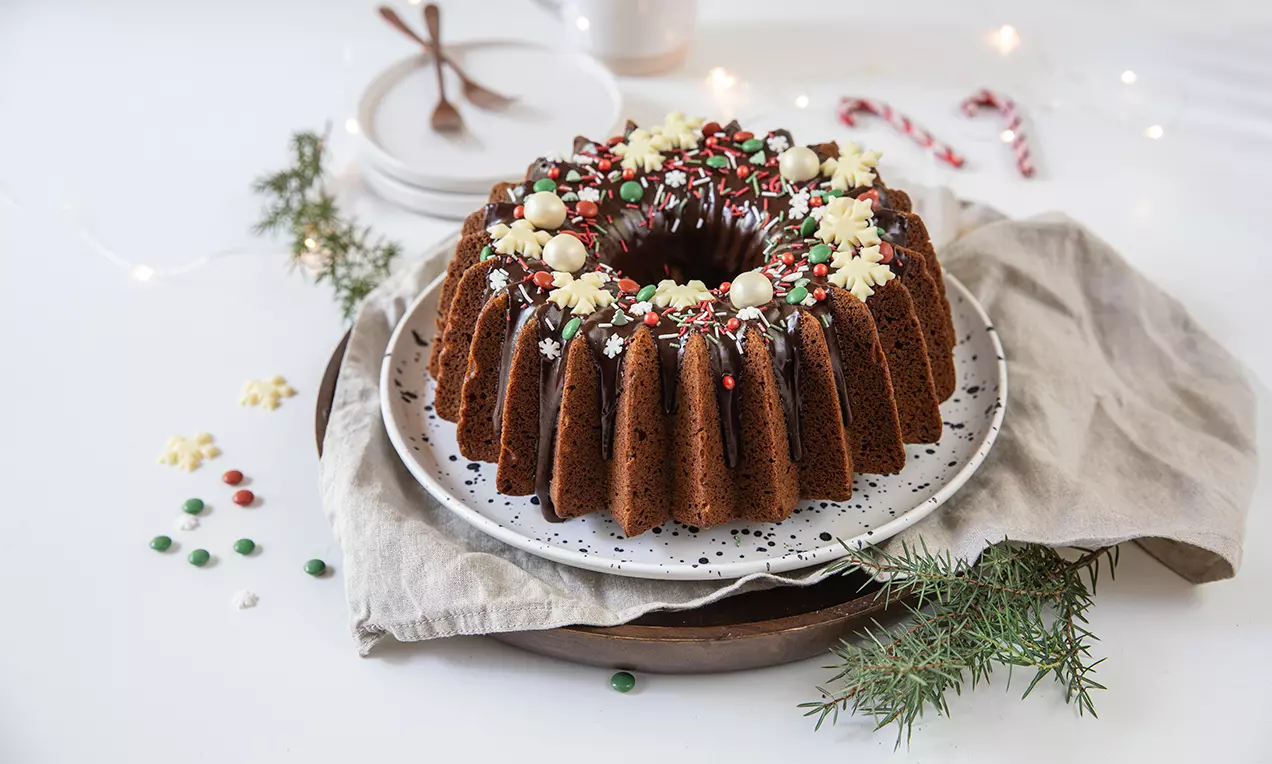 Mjuk pepparkakskrans i brun nyans med chokladglasyr, vita praliner, färgglatt strössel och gröna grankvistar på ett fat med linne servett